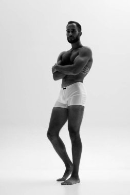 Kaslı adam beyaz boxer donlar içinde duruyor, beyaz arka plana karşı güçlü fiziğini sergiliyor. Monokrom filtresi. Güzellik ve moda kavramı, tarz ve gençlik, erkek sağlığı.