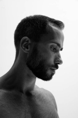 Sakallı adam beyaz arka plana karşı düşünceli bir ifadeyle profilde duruyor. Siyah beyaz filtreli fotoğraf. Güzellik ve moda kavramı, tarz ve gençlik, erkek sağlığı.