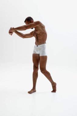 Adam dik duruyor, vücudunu hafifçe bükerken öne uzanıyor. Kasları ve güçlü bacakları beyaz arka plana karşı vurgulanmıştı. Güzellik ve moda kavramı, tarz ve gençlik, erkek sağlığı.