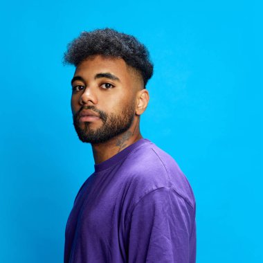 Mor gömlekli genç Afro-Amerikalı adam mavi arka planda ciddi bir ifadeyle kameraya bakıyor. İnsan duyguları, gençlik, sosyal medya, moda ve tarz kavramı.