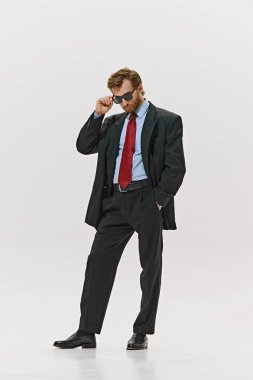 Arp giyimli adam güneş gözlüğünü ayarlıyor, kırmızı kravat takıyor ve çift göğüslü ceket giyiyor, beyaz stüdyo arka planına güvenle bakıyor. Kurumsal kültür kavramı, liderlik. Ad