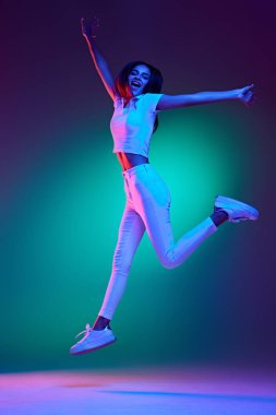 Çekici bir genç kızın uzun bir fotoğrafı, başarılı bir atlayış, el kaldırma ve neon ışıkta stüdyonun arka planına karşı bağırma. Duygular, iş, güzellik ve moda kavramı, satış. Ad