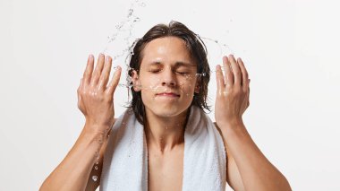 Omuzlarında havluyla yüzlerini yıkayan ve beyaz arka planda hareket eden genç ve çekici bir adamın yakın plan fotoğrafı. Doğal güzellik, moda, erkek sağlığı kavramı. Ad