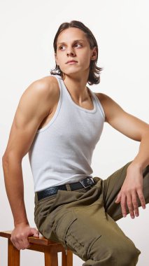 Beyaz tişört ve hacky renk pantolon giymiş genç adam özgüvenle poz veriyor, beyaz stüdyo arka planında ahşap taburede oturuyor. Doğal güzellik kavramı, moda, kozmetik ürünler, erkek sağlığı. Ad