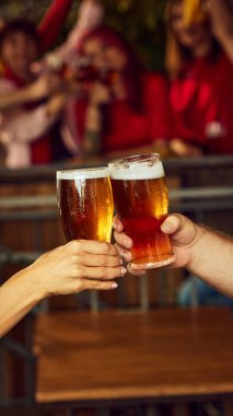 Spor hayranları arasında dinamik bir tost, bardaklar dolusu bira. Arka plan bulanıklığı paylaşılan heyecanı vurgular. Ekim festivali, bira fabrikası, tatil, cuma havası. Seçici odak.