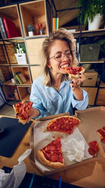 Çalışan pizza yiyor, görünüşe göre bu alışkanlığın verimliliğini nasıl etkilediğinden habersiz. Dağınık çalışma alanı kötü zaman yönetimini yansıtır. İş kavramı, insanlar ve günlük iş sorunları. Ad.