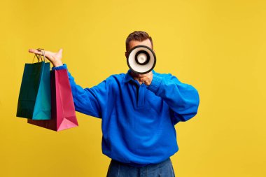 Stylish man lifting colorful bags with megaphone in hand, announcing shopping deals against sunny yellow background. Concept of discount deals, shopping spree, Black Friday, marketing promotion. clipart