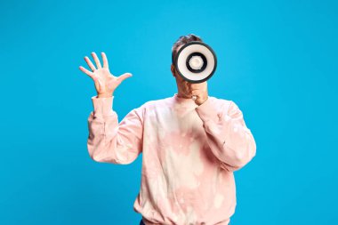 Genç adam megafonla konuşurken eliyle hareket eder, mavi arka planda dikkat çekmeye çalışır. Toplum önünde konuşma, pazarlama, duyuru, seyirci ilgisi kavramı.