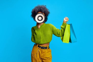 Fashionable woman in green sweater holds megaphone in one hand and shopping bags in other, signaling exciting promotion. Concept of retail promotion, Black Friday, seasonal sale, marketing. clipart