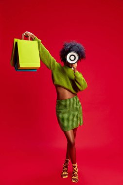 Young fashionista in green outfit raises her arm with colorful shopping bags, commanding attention with megaphone. Concept of fashion, retail, communication, announcement, sale, style clipart