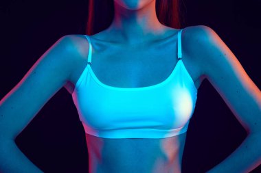 Yakın plan çekim, siyah stüdyo arka planına karşı parlak neon ışıkta vücudunun üst kısmına odaklanıyor. Mankenlerin atletik figürleri. Kadınlık kavramı, güzellik, vücut estetiği, kozmetik. Ad