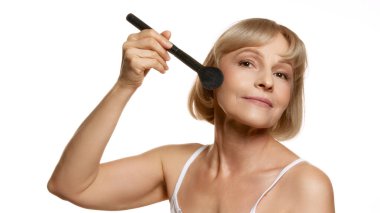 Makyaj fırçalı gülümseyen kadın, pudra sürmek üzere, beyaz stüdyo arka planıyla güzellik rutininde neşe gösteriyor. Güzellik, sağlık, kozmetoloji, yaşlanmayı önleyici ürünler. Ad