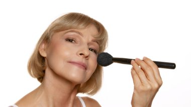 Makyaj fırçalı gülümseyen kadın, kozmetik uygulamak üzere, beyaz stüdyo arka planıyla güzellik rutininde neşe gösteriyor. Güzellik, sağlık, kozmetoloji, yaşlanmayı önleyici ürünler. Ad