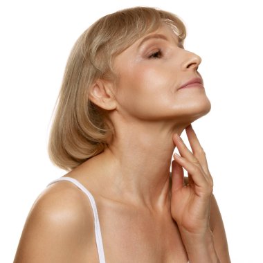 Boynunu hafifçe okşayan orta yaşlı kadın, beyaz stüdyo geçmişine karşı sakin bir ifadeyle yaşlanma zarafetini kucaklıyor. Güzellik, sağlık, kozmetoloji, yaşlanmayı önleyici ürünler. Ad