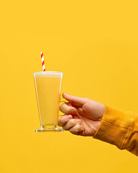 Hand in warm, knitted sweater holding glass of hot mead with red-white straw against warm yellow studio background. Concept of festive winter drink, cafe and catering, Christmas mood. Ad