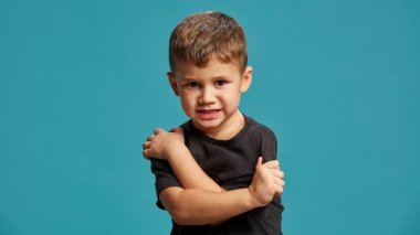 Çocuk hayal kırıklığı belirtileri gösteriyor, kollarını kavuşturmuş mavi stüdyo arka planına kararlı bir bakış atıyor. İsyan ve kendini koruma karışımı. Yüz ifadeleri, duygular. Ad