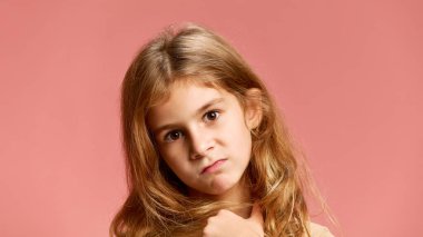 Ciddi ve yoğun bir ifadeye sahip küçük bir kız yumuşak pembe stüdyo arka planına karşı kameraya bakıyor. Meraktan. Yüz ifadeleri, olumlu ve olumsuz duygular. Ad