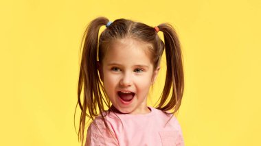 Little girl with funny pig-tails reacts with open mouth, showing surprise and excitement against sunny yellow background. Concept of facial-expressions, positive and negative emotions. Ad clipart