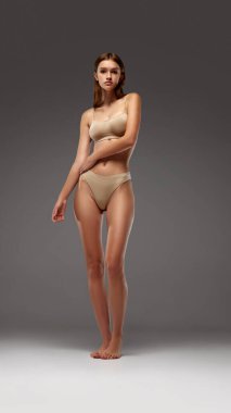 İnce, güzel bir kadın, zayıf ve zayıf bir şekilde duruyor, doğal kıvrımlarını sıcak gri arka plana vurguluyor. Kadın sağlığı diyeti kavramı, doğal güzellik, sağlık. Ad