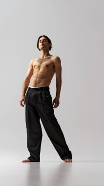 Gri stüdyo geçmişine karşı bağımsızlık ve özgüven pozu veren siyah pantolonlu genç sporcunun tam boy portresi. Doğal güzellik kavramı, erkek sağlığı, moda, cilt bakımı. Ad