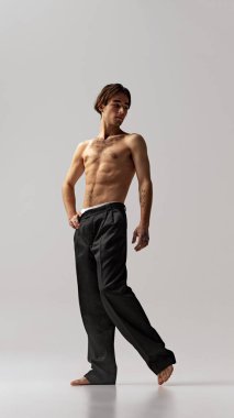 Siyah pantolonlu bir adam, hafifçe yana dönmüş, gri stüdyo arka planına karşı elini nazikçe tutarak poz veriyor. Doğal güzellik kavramı, erkek sağlığı, moda, cilt bakımı. Ad