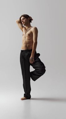 Siyah pantolonlu bir adam tek bacağı üzerinde dengede duruyor, kolları havada, gri stüdyo geçmişine karşı özgürlük ve hafiflik gösteriyor. Doğal güzellik kavramı, erkek sağlığı, moda, cilt bakımı. Ad