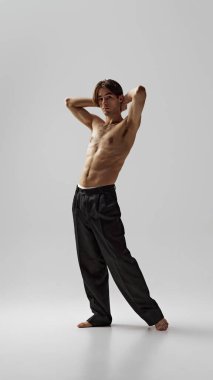 Siyah pantolonlu, kolları havada poz veren yarı çıplak genç adam gri stüdyo geçmişine karşı özgürlük ve hafiflik gösteriyor. Doğal güzellik kavramı, erkek sağlığı, moda, cilt bakımı. Ad