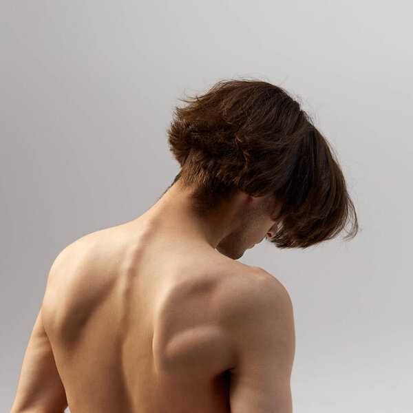 Rear view photo of young athletic man posing naked showing his healthy and muscled back against grey studio background. Concept of natural beauty, male health, fashion, skin care. Ad