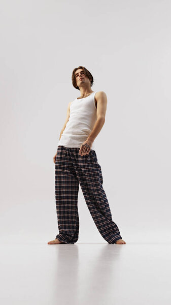 Человек в белом топе и брюках слегка откидывается назад, воплощая расслабленную позу на белом фоне студии. Концепция природной красоты, мужского здоровья, моды, ухода за кожей. Объявление