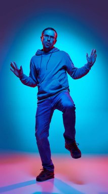 Genç adam gündelik kıyafetler giyip dans ediyor. Stüdyonun arka planına karşı neon ışıkta ellerini ve bacaklarını kaldırıyor. İnsan duyguları, güzellik ve moda kavramı, pozitif ve mizah.