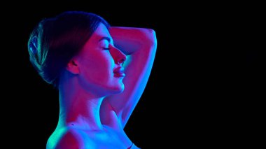 Siyah stüdyo arka planına karşı parlak neon ışıkta rahatlık ve neon ışığı yansıtan, gözleri kapalı genç, dingin bir kadının yan görüntüsü. Güzellik kavramı, kendine bakım, kozmetik tedavisi, cilt bakımı. Ad