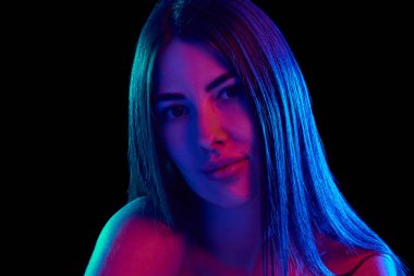 Genç bir kadının, parlak neon ışıkta siyah stüdyo arka planına karşı sakin ve kendinden emin bir ifadeyle kameraya bakışının yatay fotoğrafı. Kişisel bakım kavramı, kozmetik tedavisi, cilt bakımı. Ad