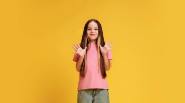 Mutlu, şaşırmış küçük kız, gündelik giyinmiş şok içinde ellerini kaldırıyor ve sarı stüdyo arka planına bakan kameraya bakıyor. Çocukluk kavramı, yaşam tarzı, ifadeler, moda. Ad