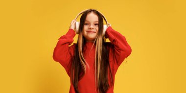 Kırmızı renkli süveter giyen, kulaklıklarını ayarlayan, sarı stüdyo arka planına bakan gülümseyen kız. Çocukluk kavramı, yaşam tarzı, ifadeler, moda. Ad