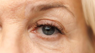 Close-up of mature womans eye, showcasing gentle wrinkles and lashes against white studio background. Thoughtful expression. Concept of natural beauty, spa treatment, cosmetology. clipart