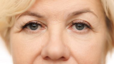 Close-up view of womans eye, emphasizing wrinkles and calm, serene gaze against white studio background. Lashes and brows. Concept of natural beauty, spa treatment, cosmetology. clipart