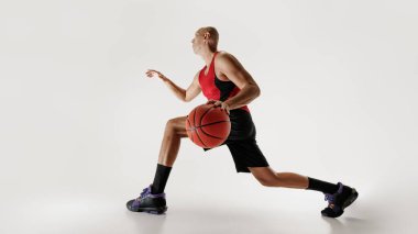 Kolsuz tişörtlü ve şortlu basketbolcu, gri stüdyo arka planına karşı bir sonraki hamlesini yapmaya hazır bir basketbol topuna odaklanıyor. Spor anlayışı, rekabet, aktif yaşam tarzı, oyun.