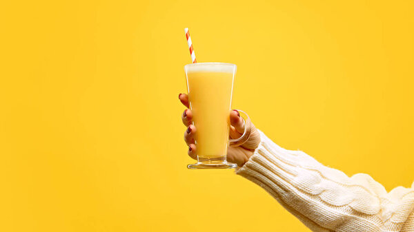 Hand in warm, knitted sweater holding glass of hot mead with red-white straw against warm yellow studio background. Concept of festive winter drink, cafe and catering, Christmas mood. Ad
