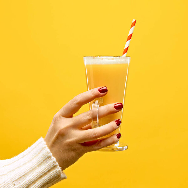 Hand in warm, knitted sweater holding glass of hot Toddy with red-white straw against warm yellow studio background. Concept of festive winter drink, cafe and catering, Christmas mood. Ad