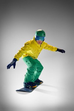 Snowboardcu, sarı ceketli ve yeşil pantolonlu genç sporcu gri stüdyo arka planına karşı havada numara yapıyor. Ekstrem spor, enerji, hız, moda kavramı.