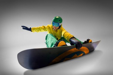 Canlı sarı ve yeşil kıyafetli snowboard sporcusu snowboard yaparken dengesini koruyor, macera ve beceriyi temsil eden gri arka plana karşı. Ekstrem spor, enerji, moda kavramı.