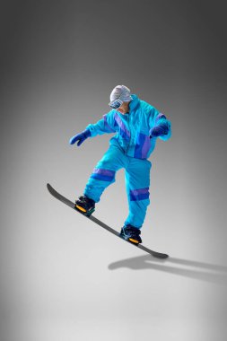 Snowboardcu, gri stüdyo arka planına karşı kalın vitesli hareketler sergiliyor. Kış sporları ve turizm konseptleri için mükemmel. Ekstrem spor, enerji, hız, moda kavramı.