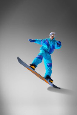 Canlı snowboardcu, gri stüdyonun arka planında kış sporlarının ve açık hava keşiflerinin ruhunu ön plana çıkarırken görüntülendi. Ekstrem spor, enerji, hız, moda kavramı.