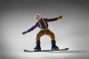 Örgü eldivenli ve pantolon askısı takmış bir adam snowboard numarasına hazırlanıyor. Gri stüdyo arka planına el uzatıyorlar. Ekstrem spor, enerji, hız, moda kavramı.