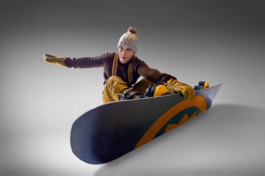 Kış vitesli bir adam, gri stüdyo arka planına karşı dinamik hareket ve kontrol gösterisi yaparak snowboard 'unda dengede durur. Ekstrem spor kavramı, enerji, hız, moda, klasik.