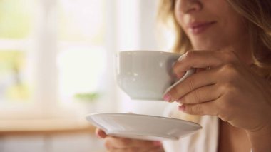 Beyaz kahve fincanı ve çay tabağı tutan güzel, genç bir kadının yakın çekimi. Yumuşak, aydınlık manzaralı. Uyanma kavramı, güzellik, hafta sonu, sabah rutini. Ad