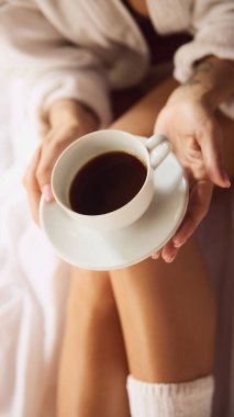 Beyaz bornozlu bir kadın, yumuşak bir battaniyeyle yatakta rahatça otururken elinde bir fincan kahve tutuyordu. Doğal ışık rahat yatak odasını aydınlatır. Uyanma kavramı, güzellik, hafta sonu, sabah rutini.