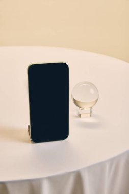 Beyaz masada şeffaf kristal kürenin yanında dikey duran akıllı telefon, iş temaları için minimum ve çok yönlü bir model sunuyor. İş kavramı, pazarlama, estetik, teşvik.