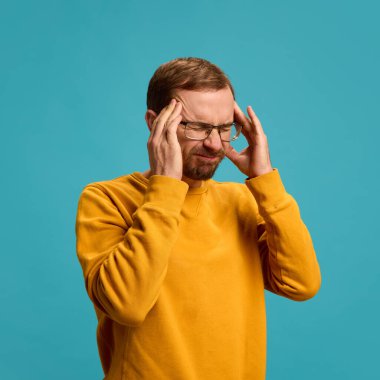 Rastgele poz veren genç bir adamın portresi, acı çekerken el ele tutuşması, turkuaz arka planda baş ağrısı. Soğuk algınlığı, virüs mevsimi, sağlık, sağlık sigortası kavramı.