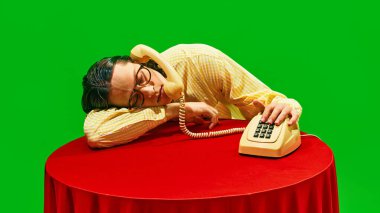 Genç adam, telefon ahizesini tutarak başını masaya yaslıyor, eski ofis ortamında yorgunluk ve bıkkınlık gösteriyor. İş kavramı, iş hayatı dengesi. Pop sanat renkleri. Nostalji.